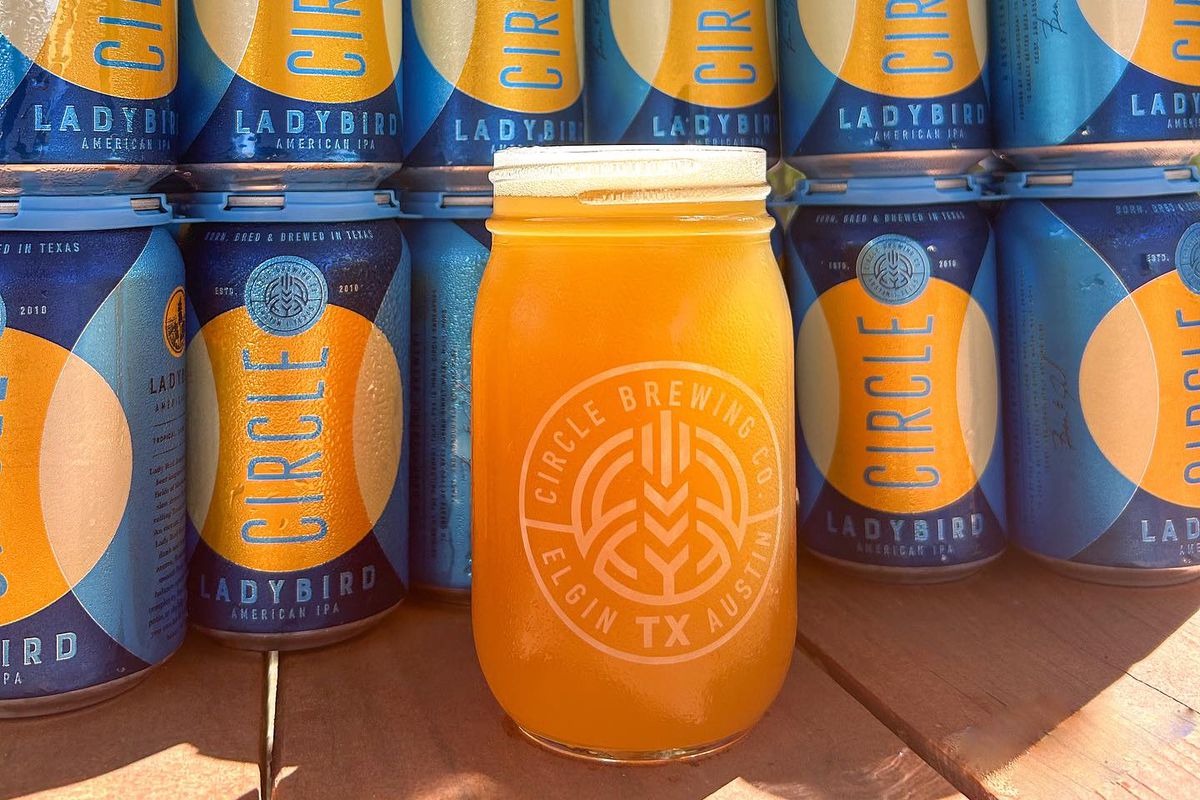 A golden beer in a mason jar in front of blue-orange cans of beers.