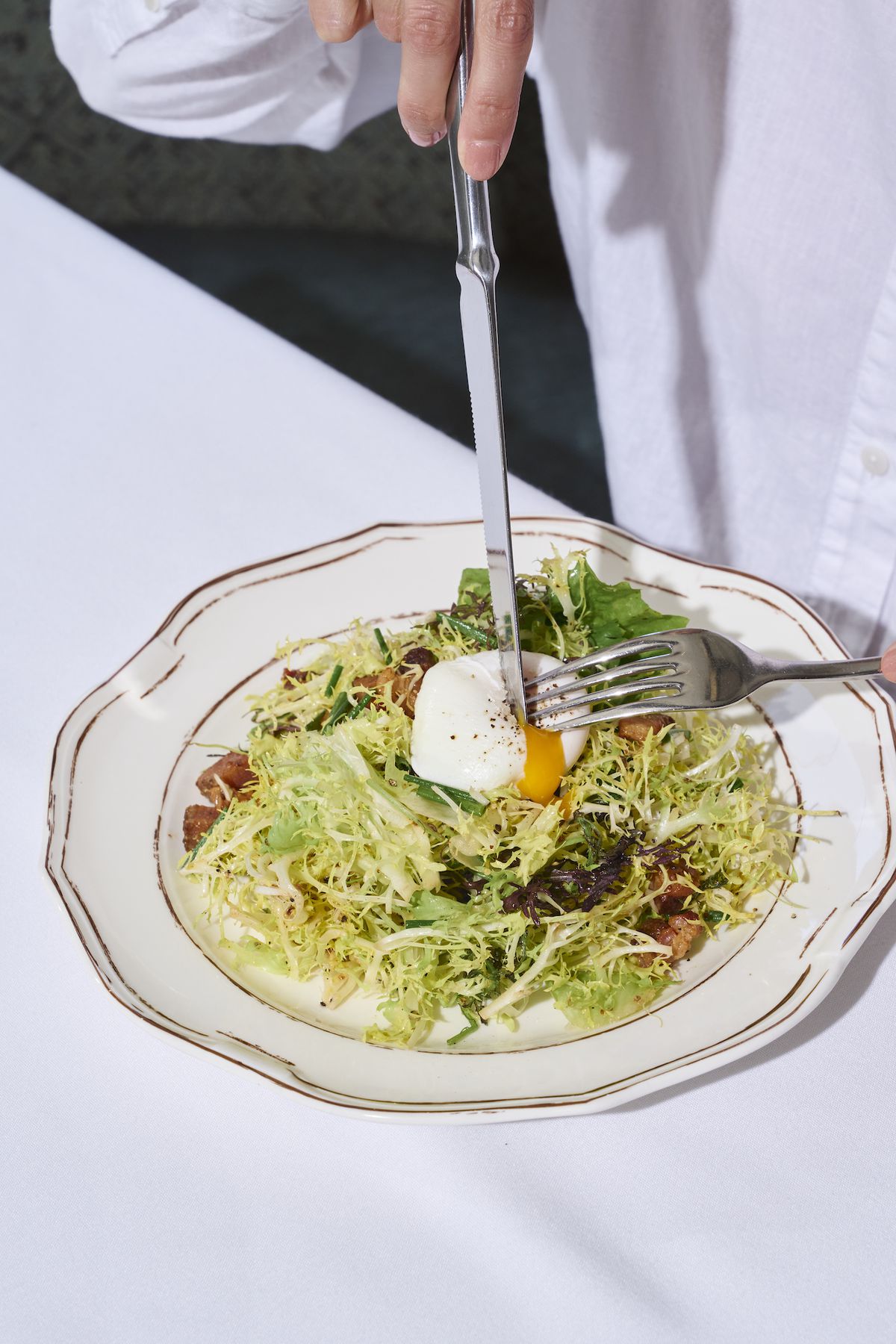 A frisee salad with soft egg with a knife going through it as a person in a white shirt cuts, inside a new restaurant Shirley Brasserie in Hollywood.