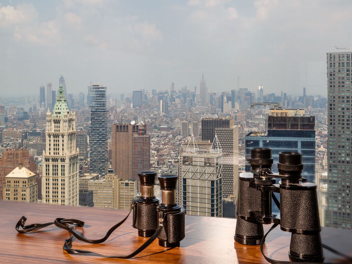 A pair of binoculars sits on a wooden table in front of views of the NYC skyline.