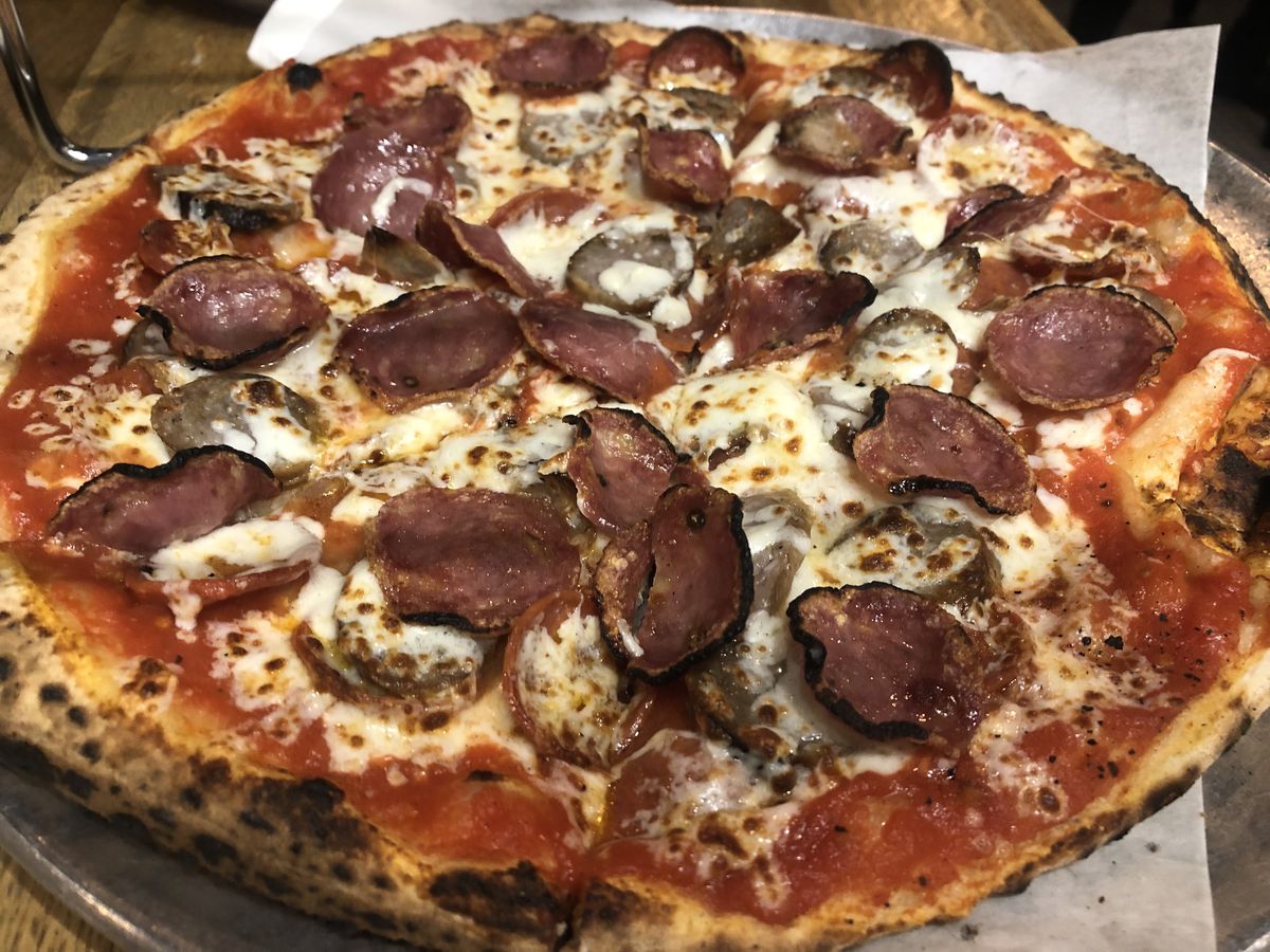 A pizza with a charred crust topped with sausage, pepperoni, soppressata, provolone, mozzarella, and a red sauce.