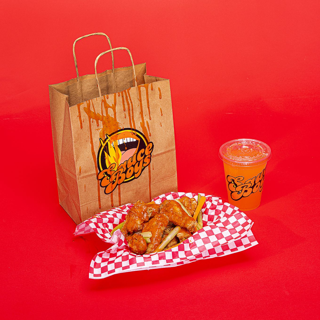 A bag with a sticker for a fake brand called Sauce Boys next to a basket of chicken wings and a clear plastic cup with another sticker for  Sauce Boys on a red backdrop.