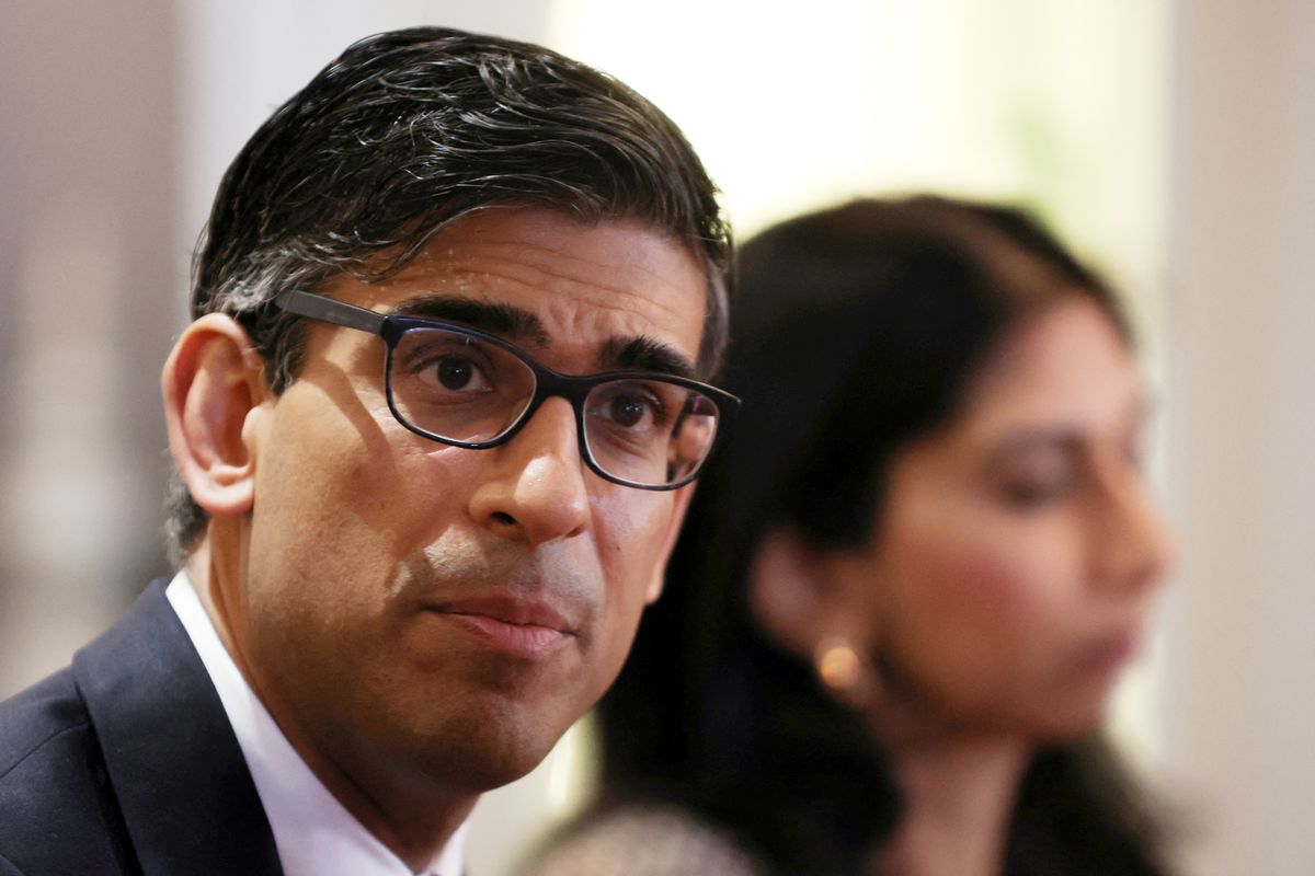 Rishi Sunak wears black rimmed glasses and a thoughtful expression. Behind him, the blurry profile of Suella Braverman is visible. 