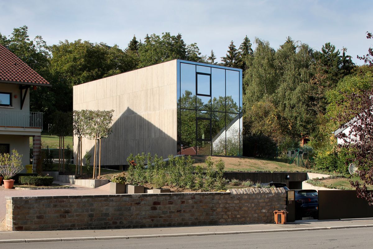 The exterior of a house which has concrete walls and a front facade which consist entirely of glass. The house is surrounded by trees.