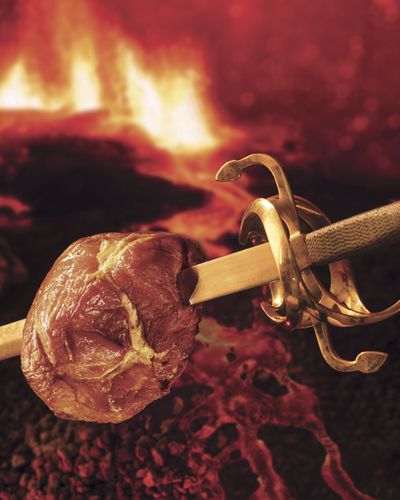 A ham impaled on a sword with a twin-serpent basket hilt, in front of a fire.