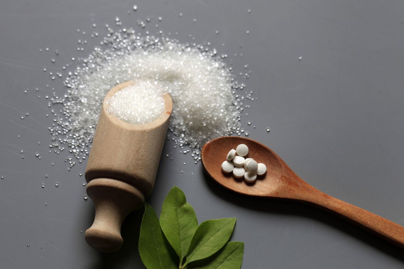 A picture of aspartame tablets in a wooden spoon and a container laying on its side with sugar spilled around it.