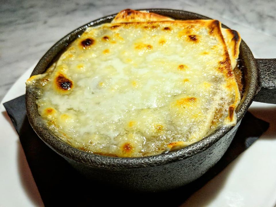 A black cast iron bowl of French onion soup, with a sheet of bubbly browned cheese across the top