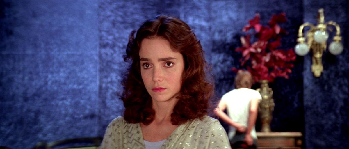 Jessica Harper in Suspiria, backgrounded by a blue wall, red flowers, and a man in a white shirt.