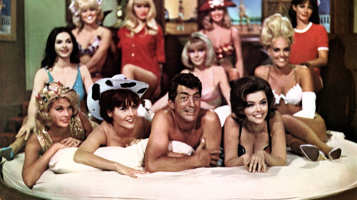 Matt Helm (Dean Martin) sits naked on a bed surrounded by women in lingerie in The Silencers