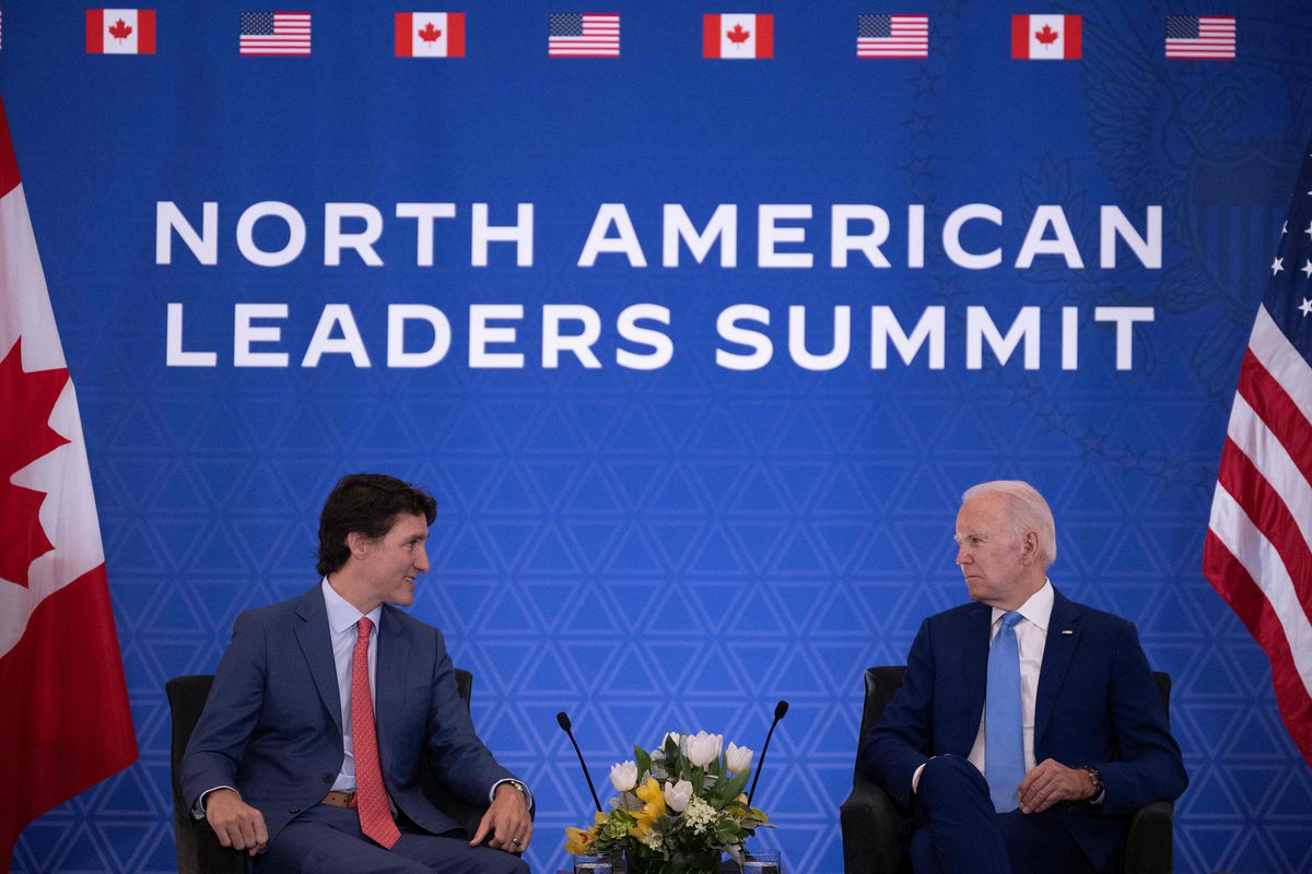 Trudeau (left) and Biden (right) are seated in front of a large blue banner with the words North American Leaders Summit written in white. Biden is seated next to an American flag. Trudeau is seated next to a Canadian flag.