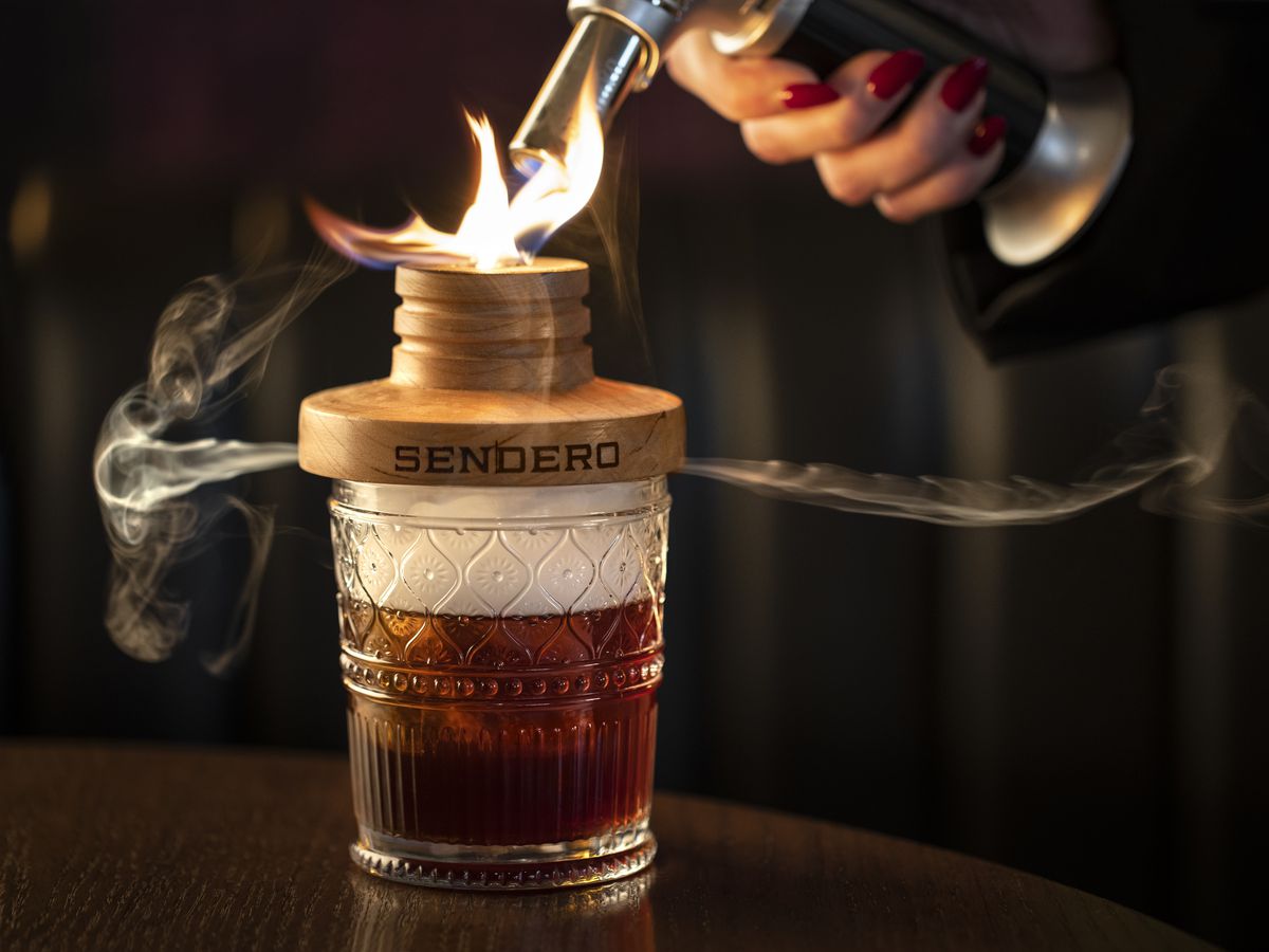 An amber cocktail in a clear glass plus a wooden smoker being smoked with palo santo.