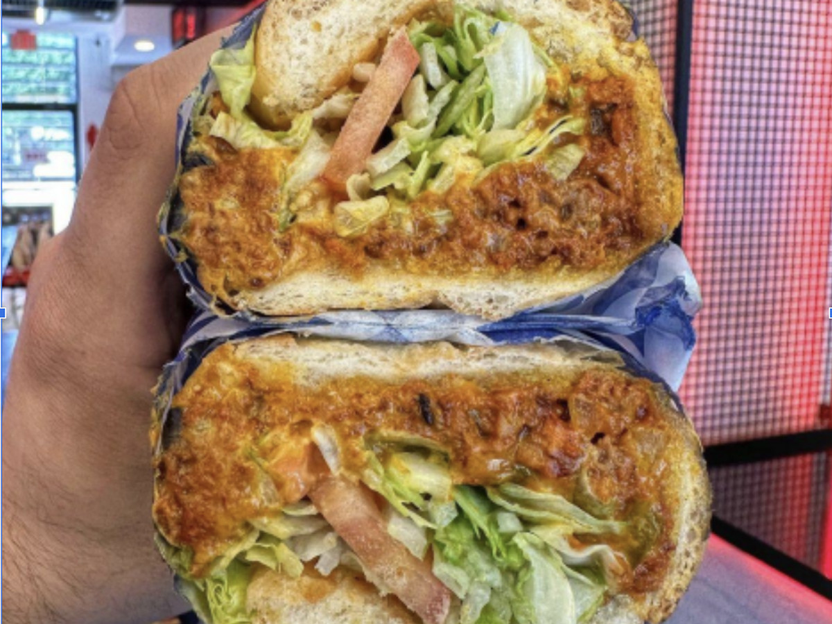 A person holds a chopped cheese hoagie filled with cheesy diced meat, bell peppers, and topped with lettuce and tomato.