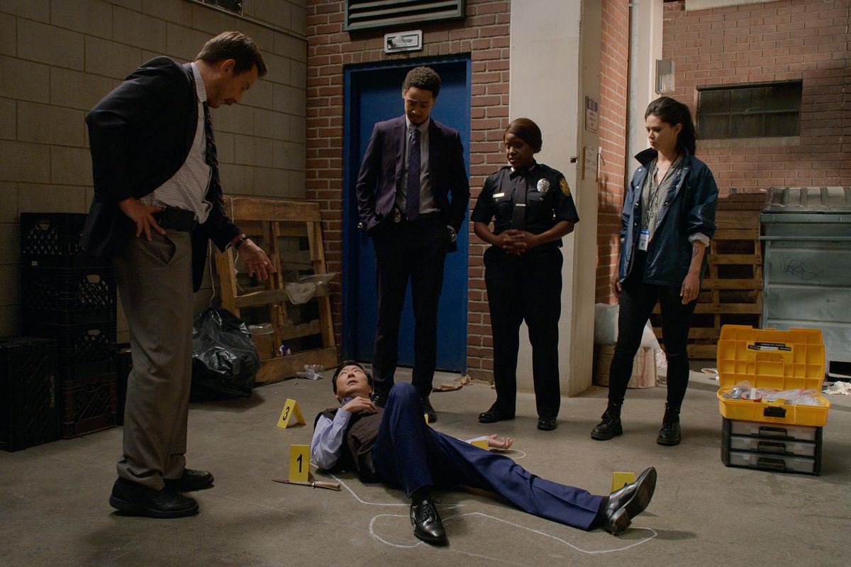 Ken Jeong lying in the place of a body while a group of detectives stand around him recreating the crime scene from the Murderville season 1 finale