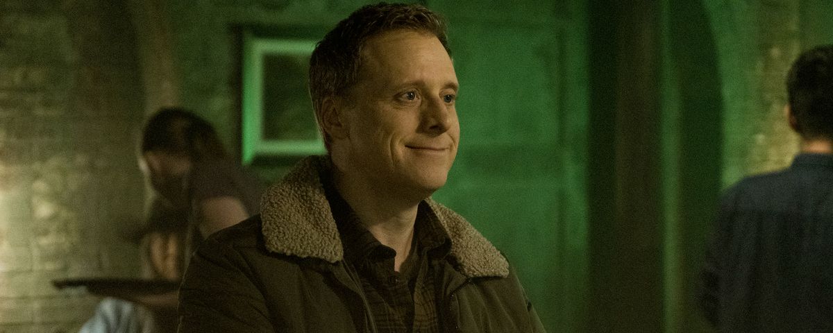 Alan Tudyk sits as a bar wearing a weird smile as a disguised alien in Resident Alien