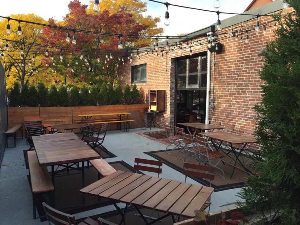 A small restaurant patio, set up against a red brick building, features wooden tables, string lights, and trees lining the edge.