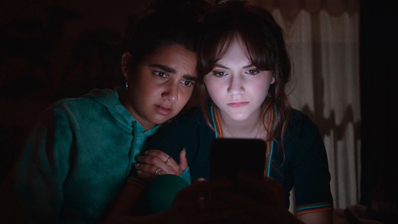 Two young women sit in the dark looking at the brightly lit screen of a phone.