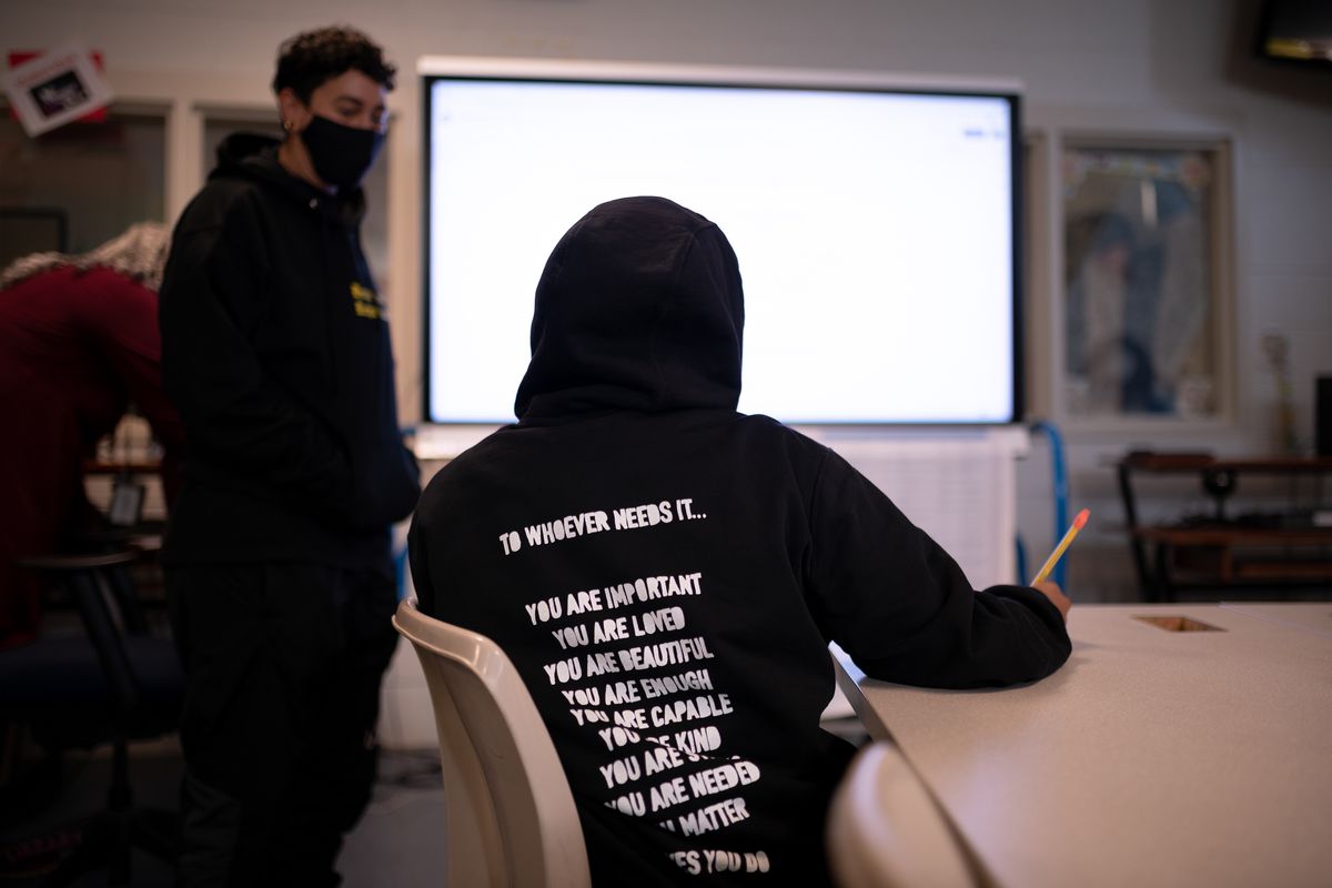 A teacher speaks with a student in the classroom, each wearing black hoodies as a screen is brightly lit behind them.