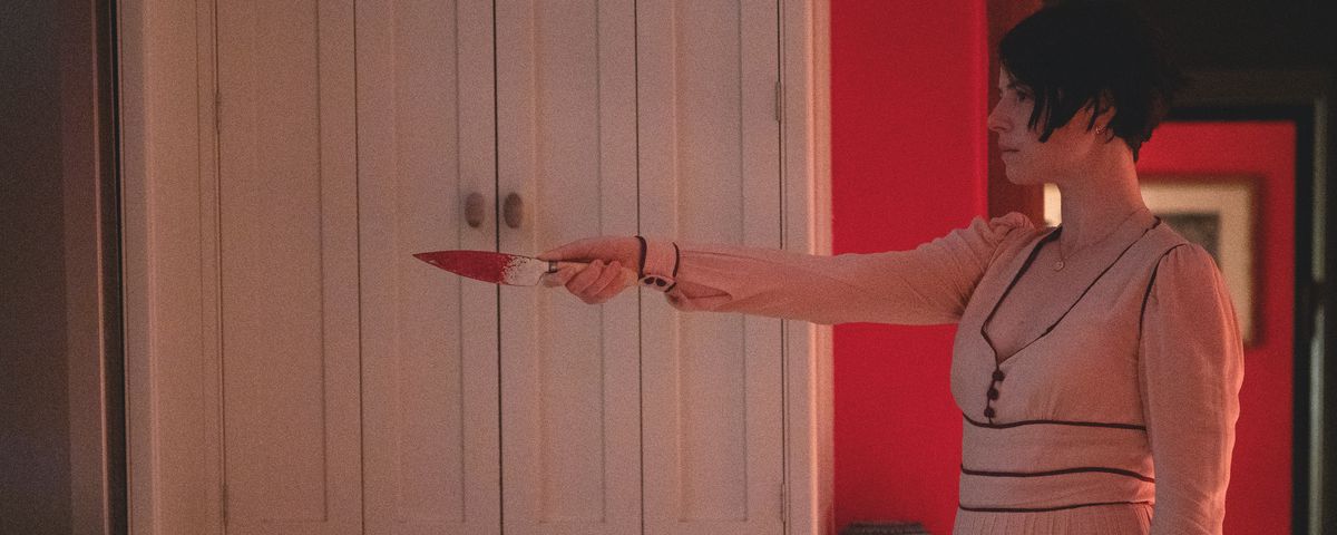 Jessie Buckley in a long pink dress, holding up a bloody knife in Men