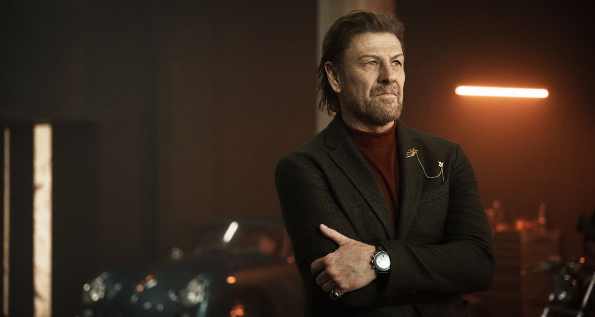 Alman Kido (Sean Bean), a pensive-looking man in a suit, stands in a dimly lit garage full of shiny, expensive-looking cars and stares vaguely off screen in Knights of the Zodiac