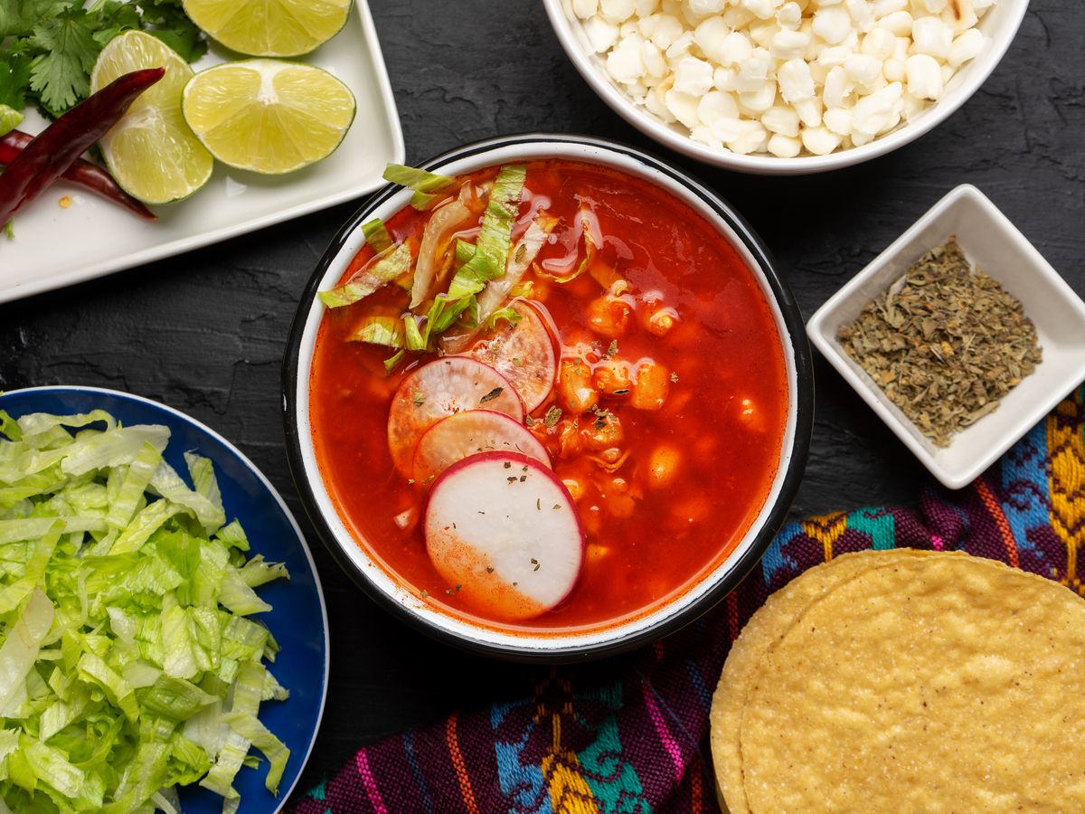 A bowl of red pozole surrounded by plates of toppings.