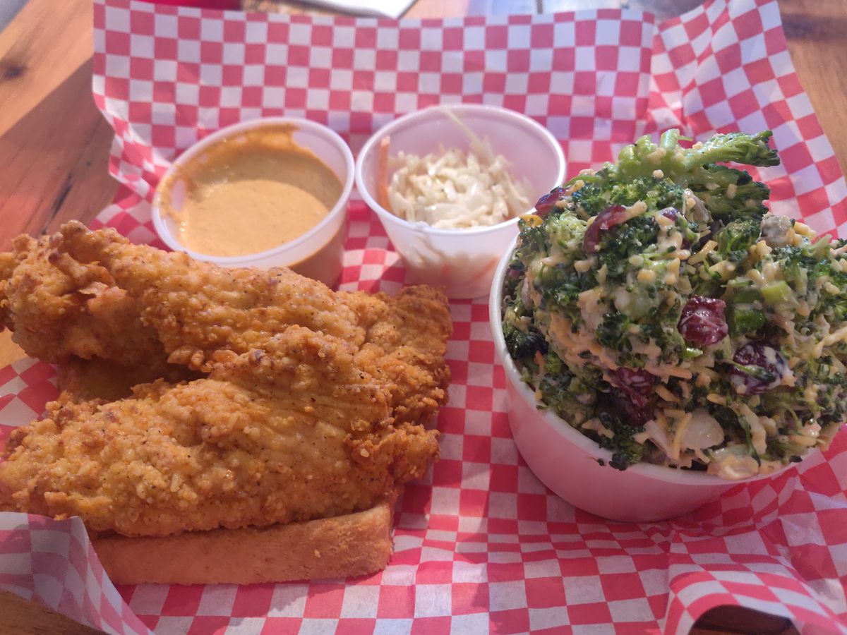 Chicken tenders, broccoli salad, and two cups of dipping sauces arranged on a red and white checkered paper.