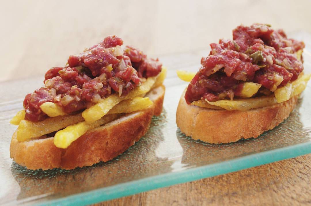 Slices of bread topped with french fries and steak tartare, arranged on a glass plate