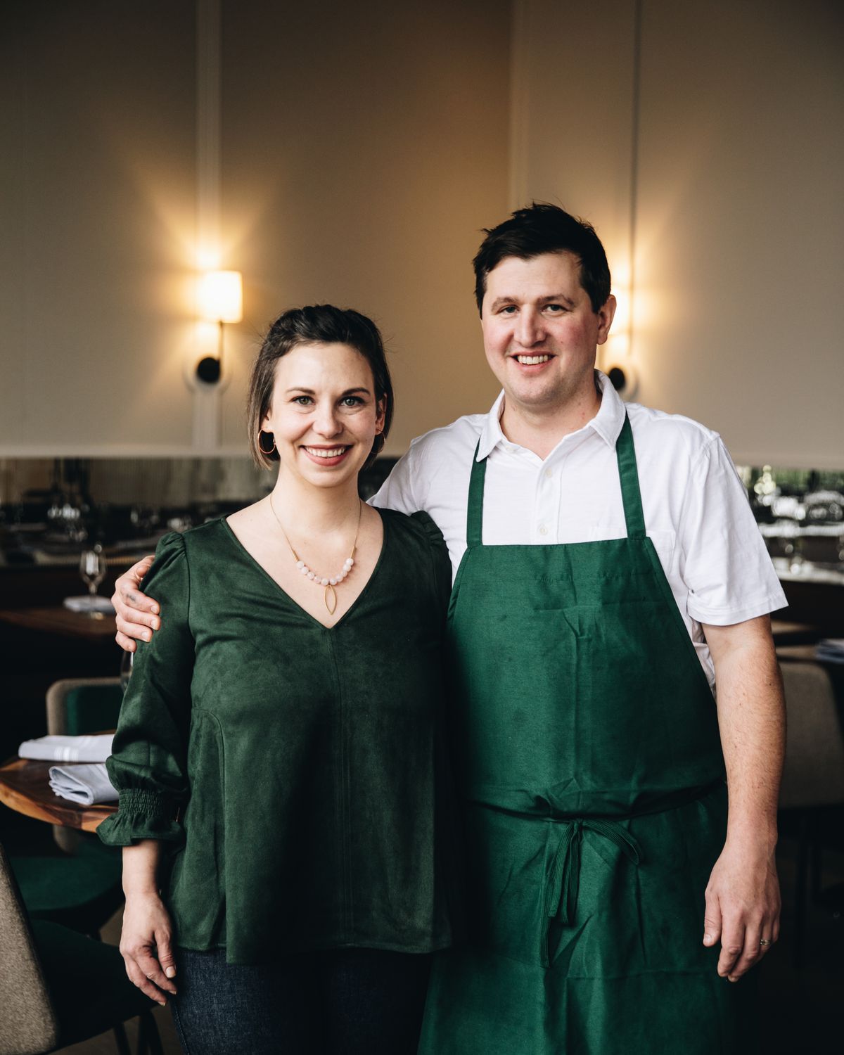 Jeanie Ritter, wearing a dark green blouse, and Adam Ritter, wearing a white shirt and dark green apron, standing in the interior of their restaurant. 
