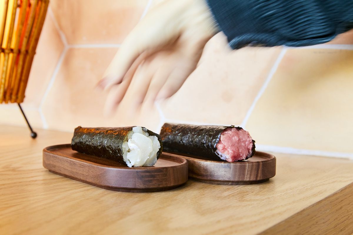 A hand blurs in to grab one of two sushi hand rolls on a wooden plate.