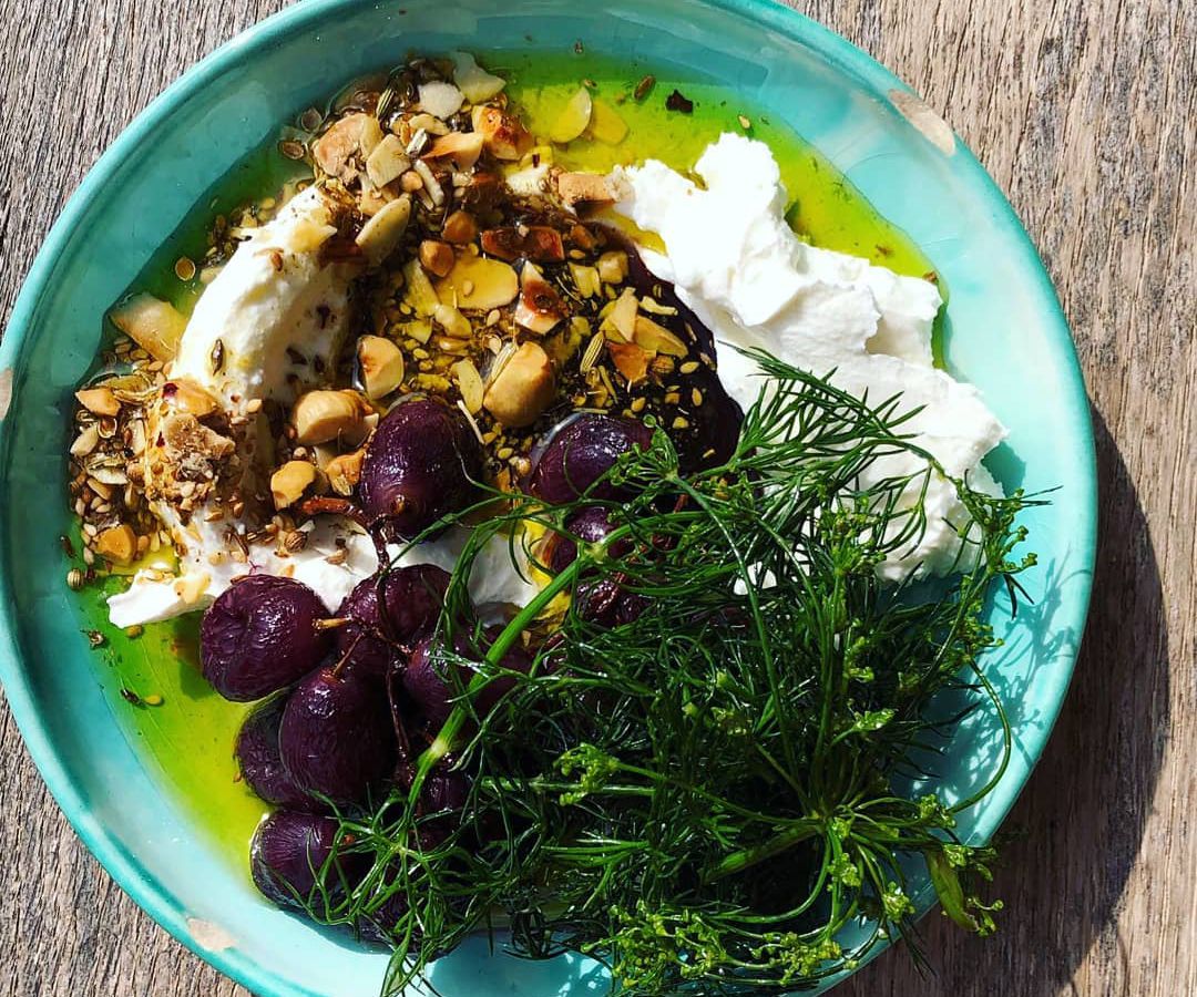 From above, a bright ceramic plate topped with a large heap of labneh topped with dukkah, a pile of dark grapes, and a bunch of greens