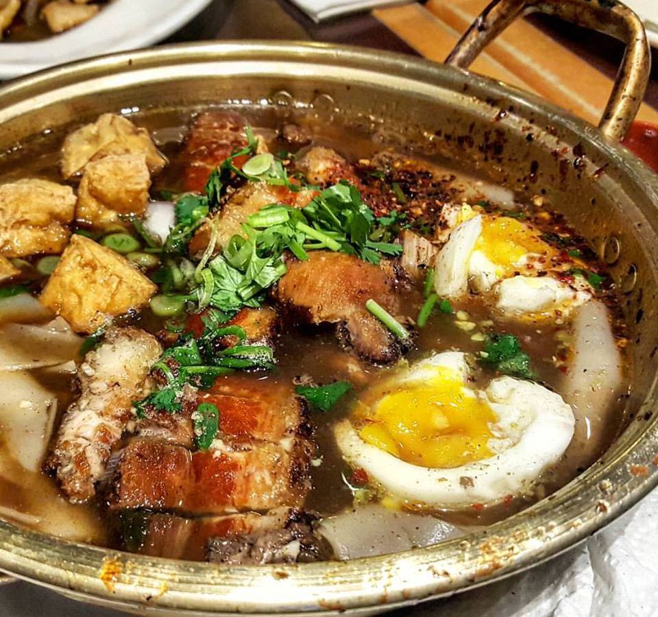 A golden bowl full of a Thai soup with a boiled egg, crispy pork, fried tofu, and herbs