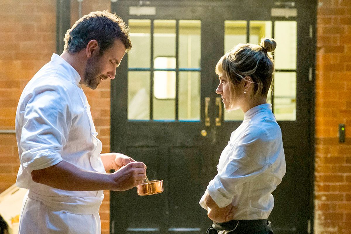 In a still from “Burnt,” Bradley Cooper — holding a copper saucier pan — and Sienna Miller wear white chef coats and face each other in front of black double doors and a brick wall.