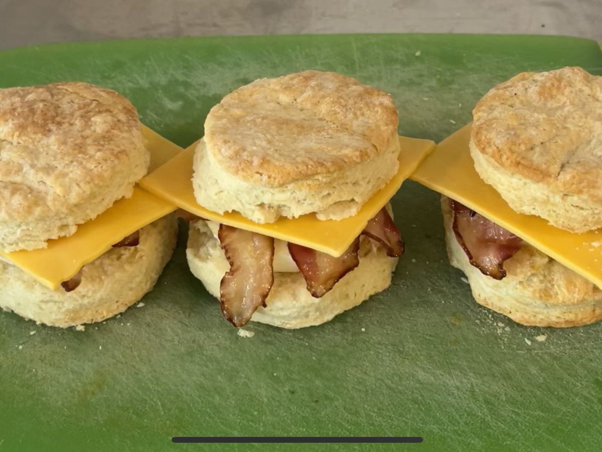 Three biscuit sandwiches with cheese, bacon, and eggs on a green board.