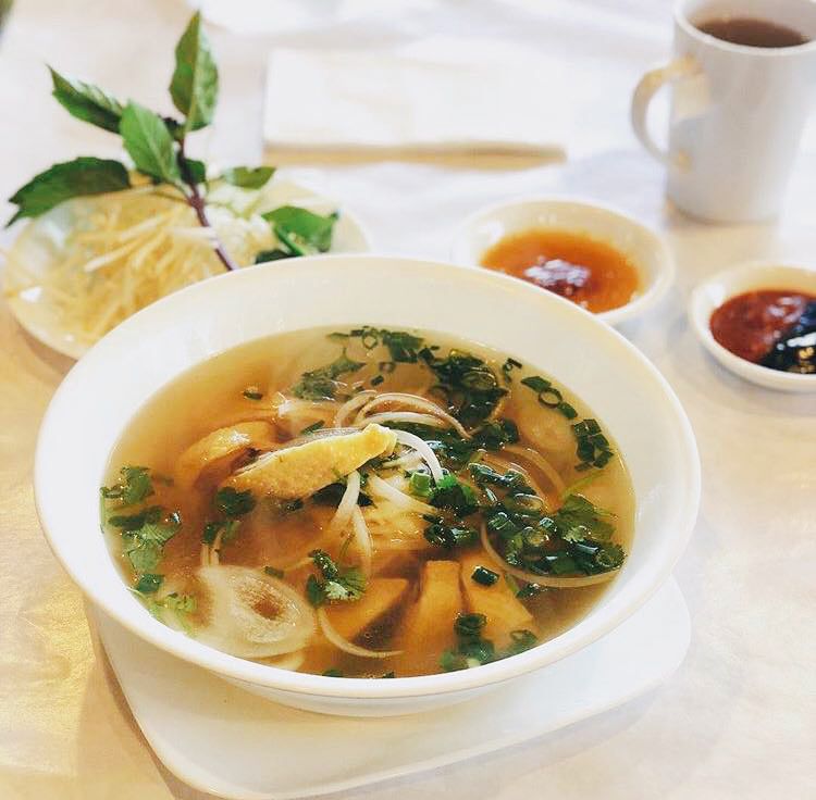 A white bowl with noodles and soup in front of a plate of bean sprouts and basil.