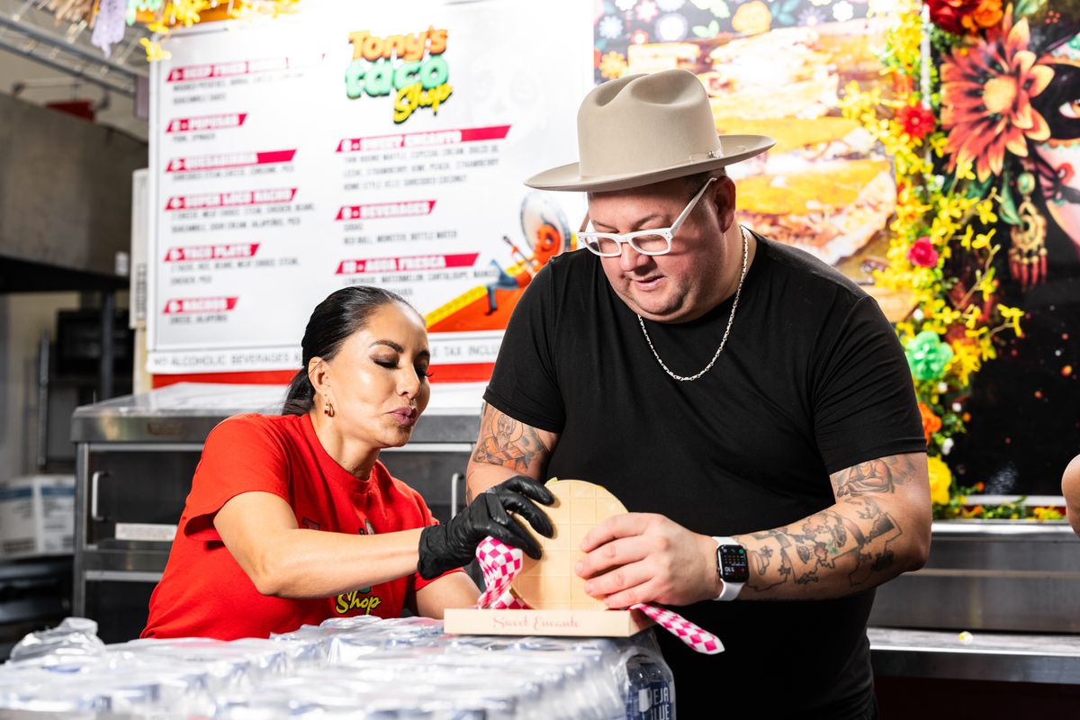 A woman helps celebrity chef Graham Elliot as he tries to get a hold of a dessert at the state fair. They both stand behind the counter at a taco stand at the state fair.