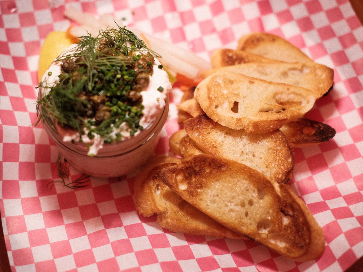 A jar of smoked trout dip topped with dill with slices of toasted baguette on a red and white checked paper.