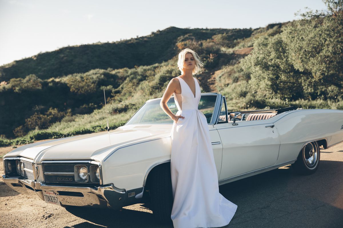 A model in a Floravere wedding gown posing near a car