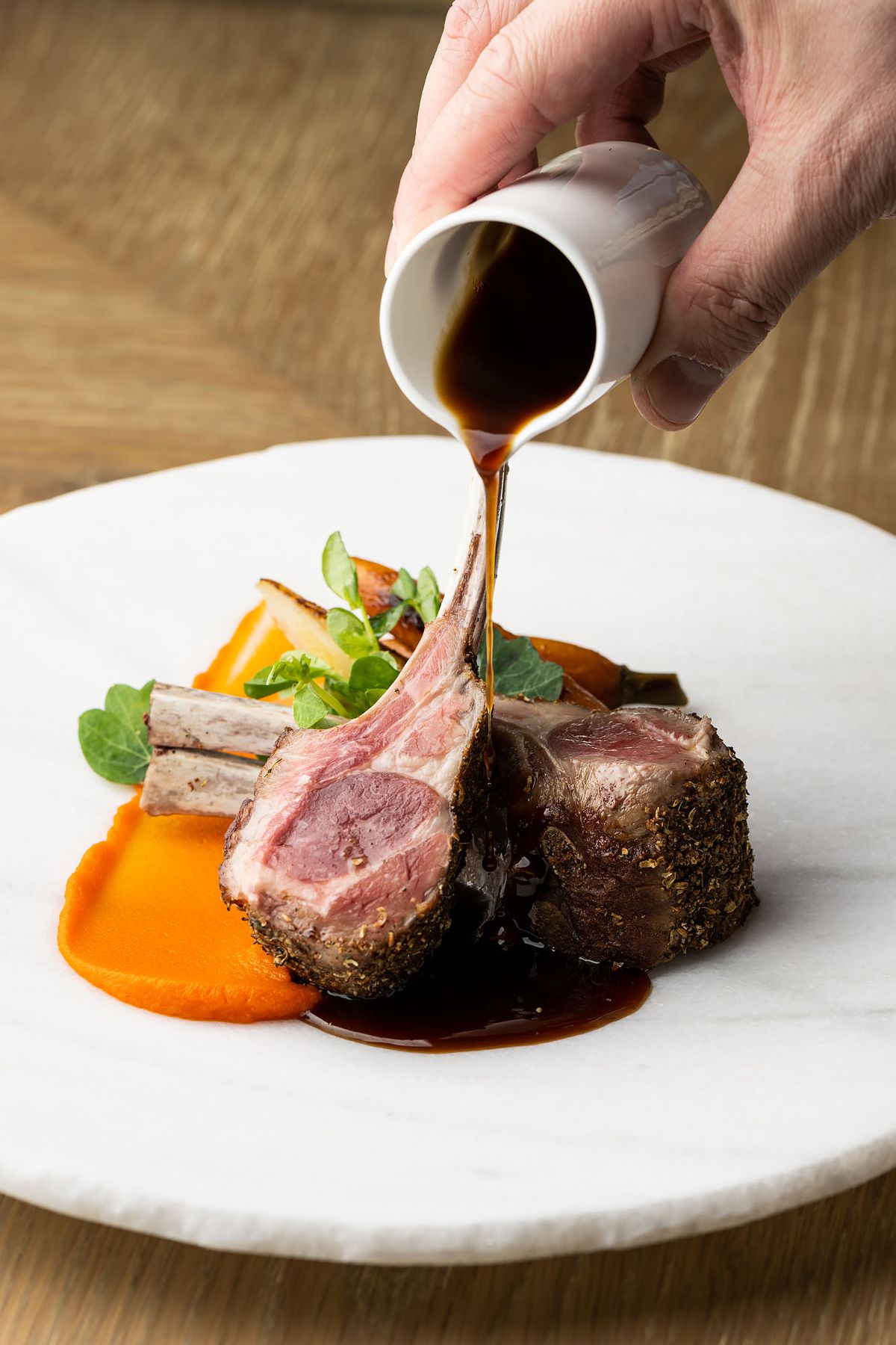 A hand pours a dark brown liquid overtop of rare-cooked lamb chops.
