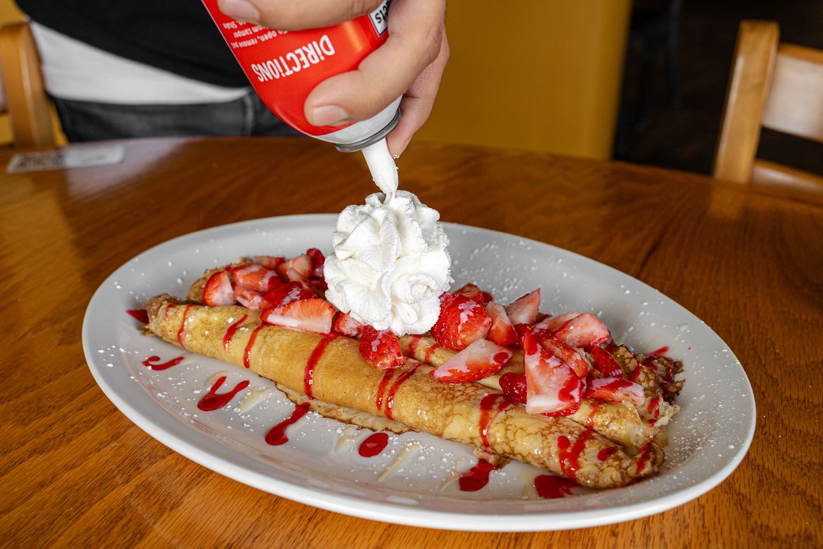 A person squeezes whipped cream from a can onto a crepe topped with strawberries.
