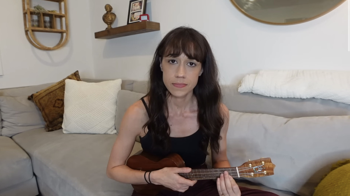 A still of Colleen Ballinger holding a ukulele in her apology song video. She is sitting on a couch and looks deeply into the camera.