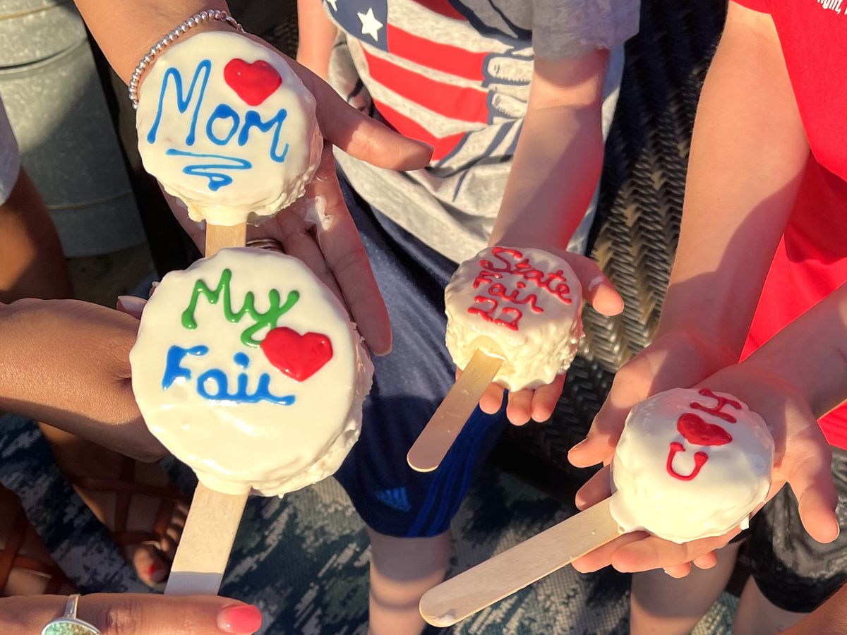 Five hands hold out small white frosted cakes on sticks with icing lettering that reads “Mom” and “My Fair” and “State Fair 22.”
