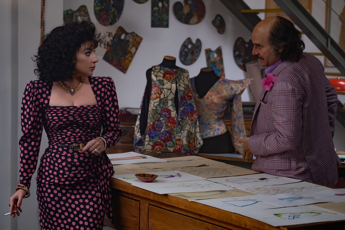 A woman in a tight square-necked polka dot dress and a bald man in a brightly colored plaid jacket with a flower on the lapel stand across a table from one another, looking at drawings of clothing.