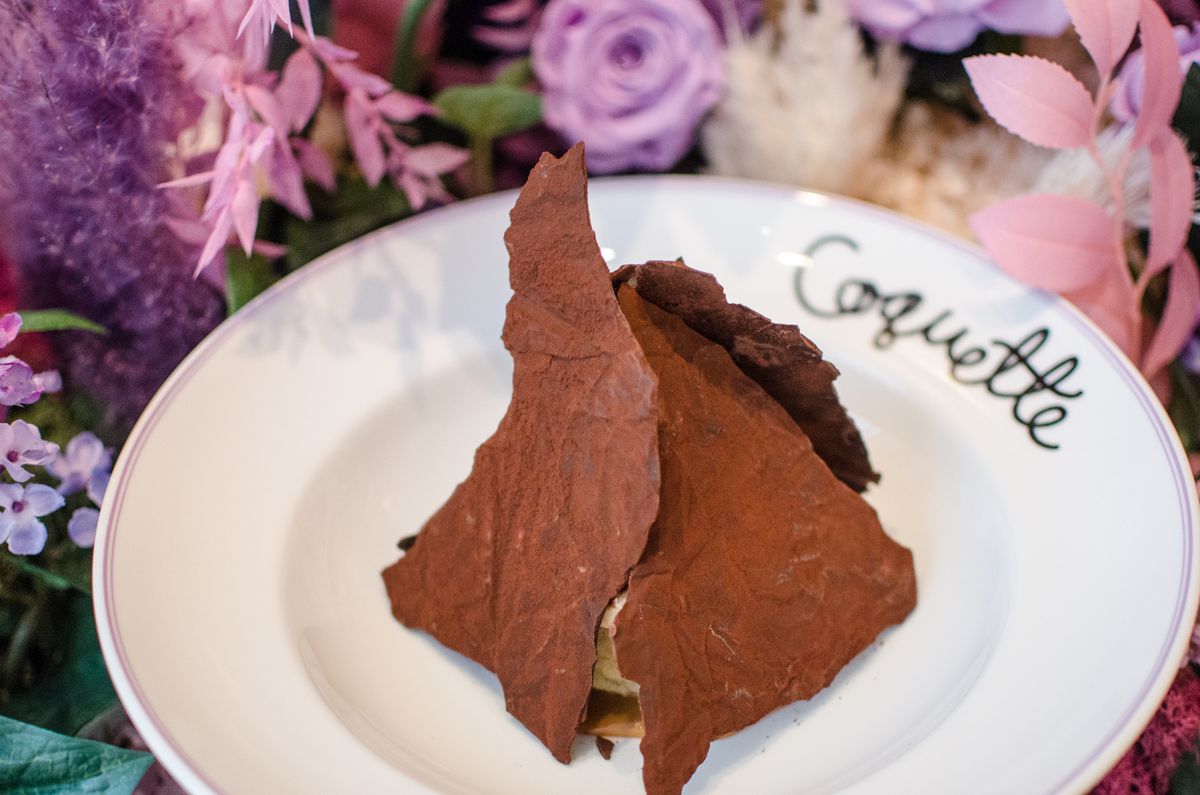 A dessert featuring three chocolate panels in a pyramid shape sits on a white plate with a backdrop of pink and purple flowers.