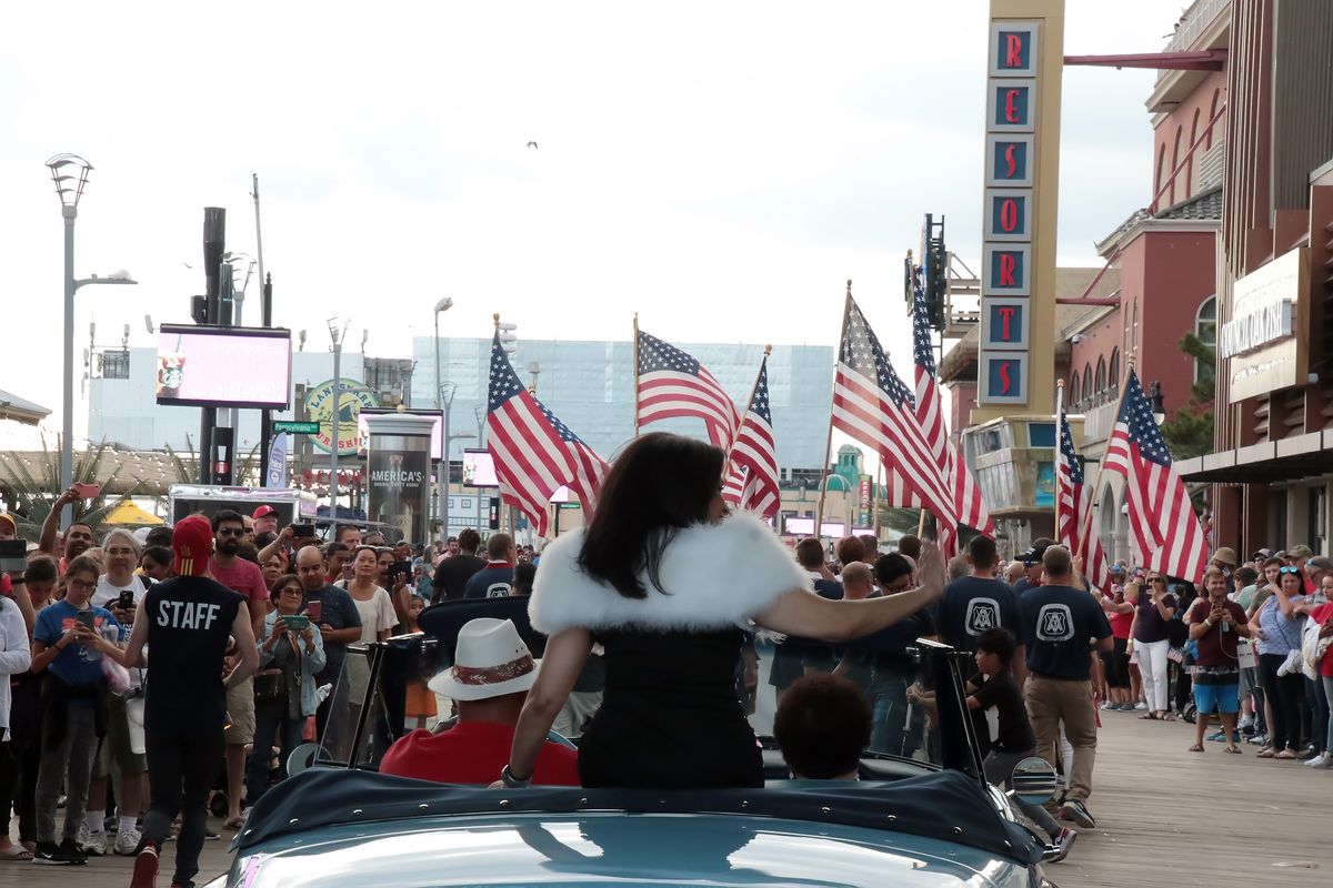 A woman sitting on the back of a convertible waves to a crowd in a parade. Many people are waving American flags.