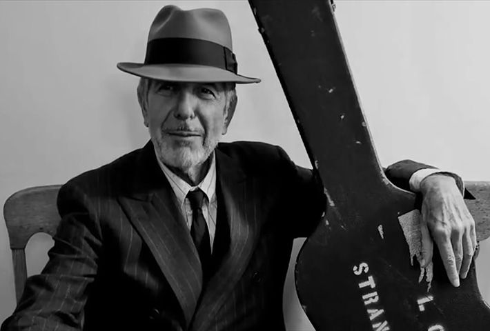 Leonard Cohen, dressed in a suit and tie and a nice hat, holds a guitar case.