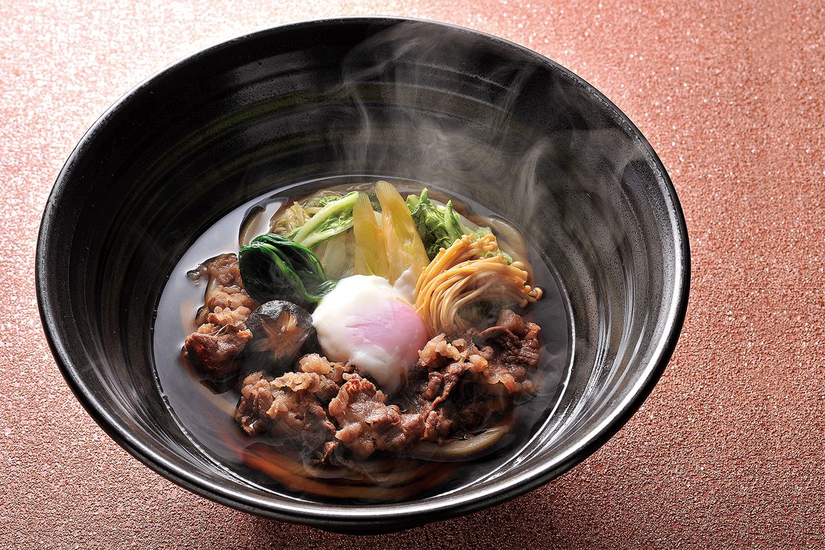 Bowl of a Japanese soup, featuring a transparent brown broth, thinly sliced beef, thick udon noodles, a soft-boiled egg, and vegetables
