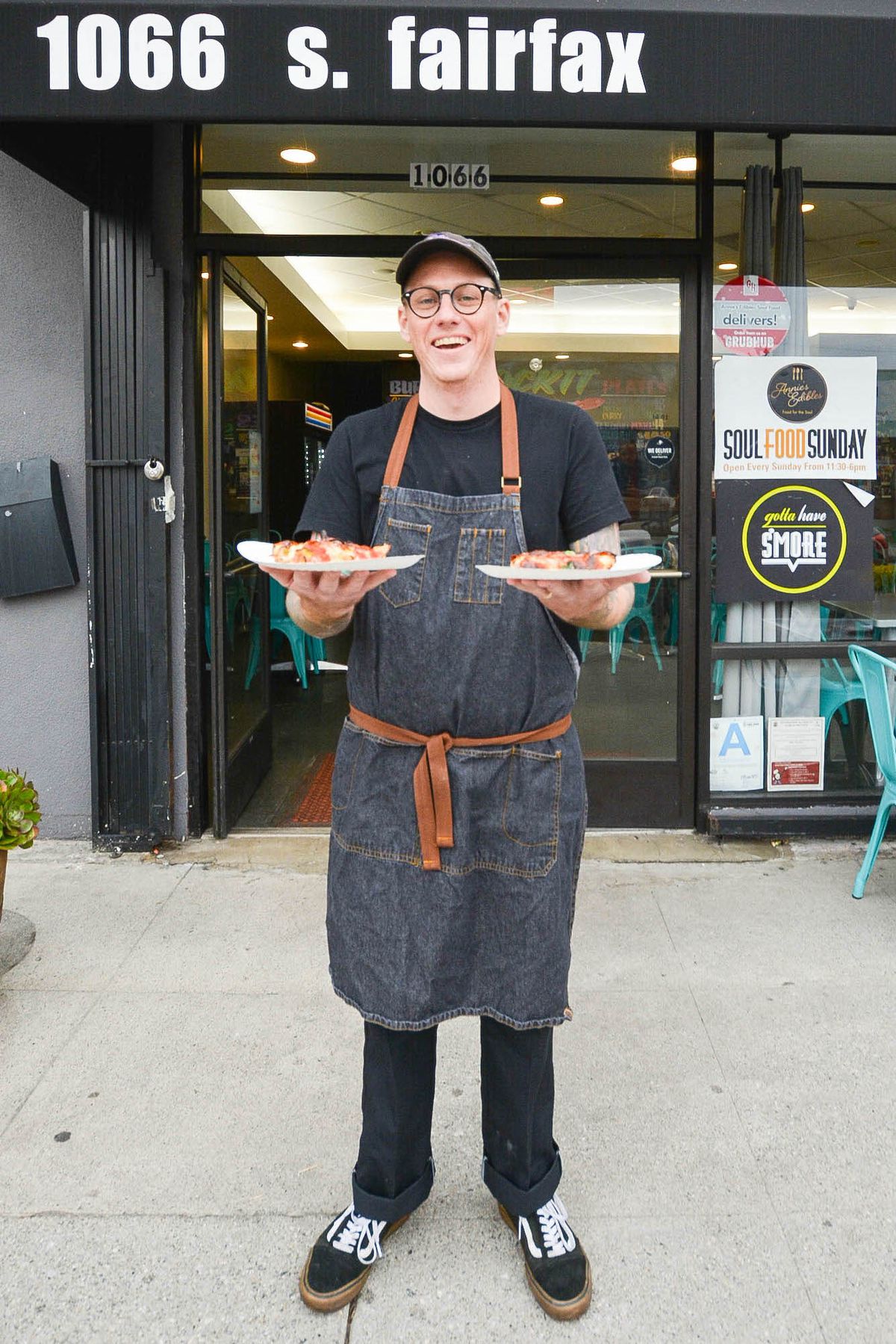 A man in a grey apron holds two slices of pizza on white paper plates in front of a restaurant.