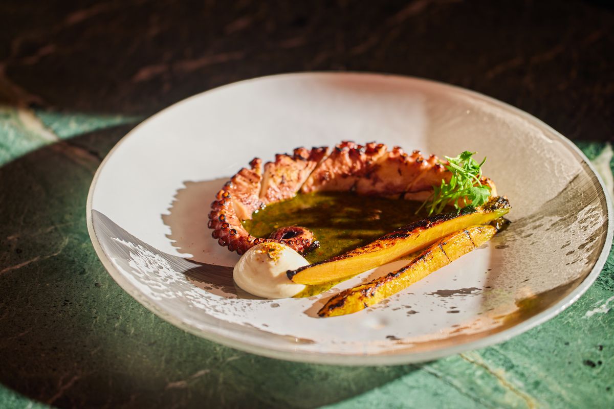 A round plate with grilled octopus, a dollop of white sauce, carrots, and garnishes.