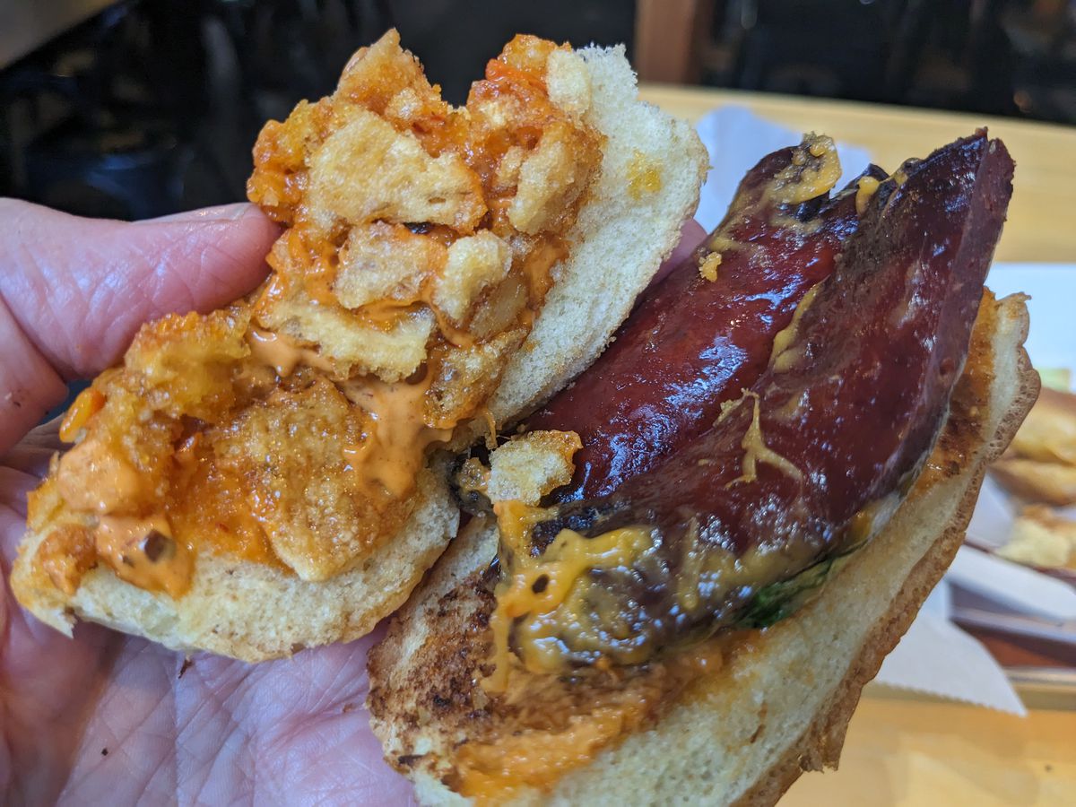 A dark red sausage split on a bun with tidbits of pork skin and and orange sauce.