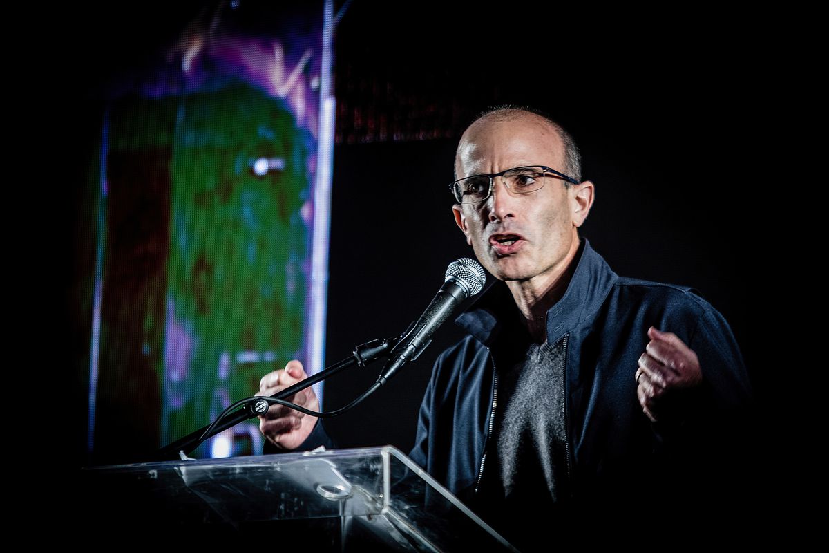 Yuval Harari stands at a lectern speaking into a microphone and gesturing with his hands. 