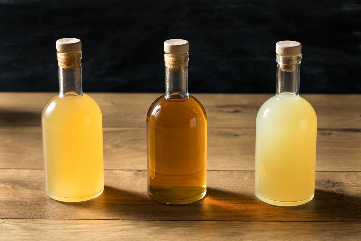 Three glass bottles of beverages, one yellow, one amber, and another yellow, on a wooden table