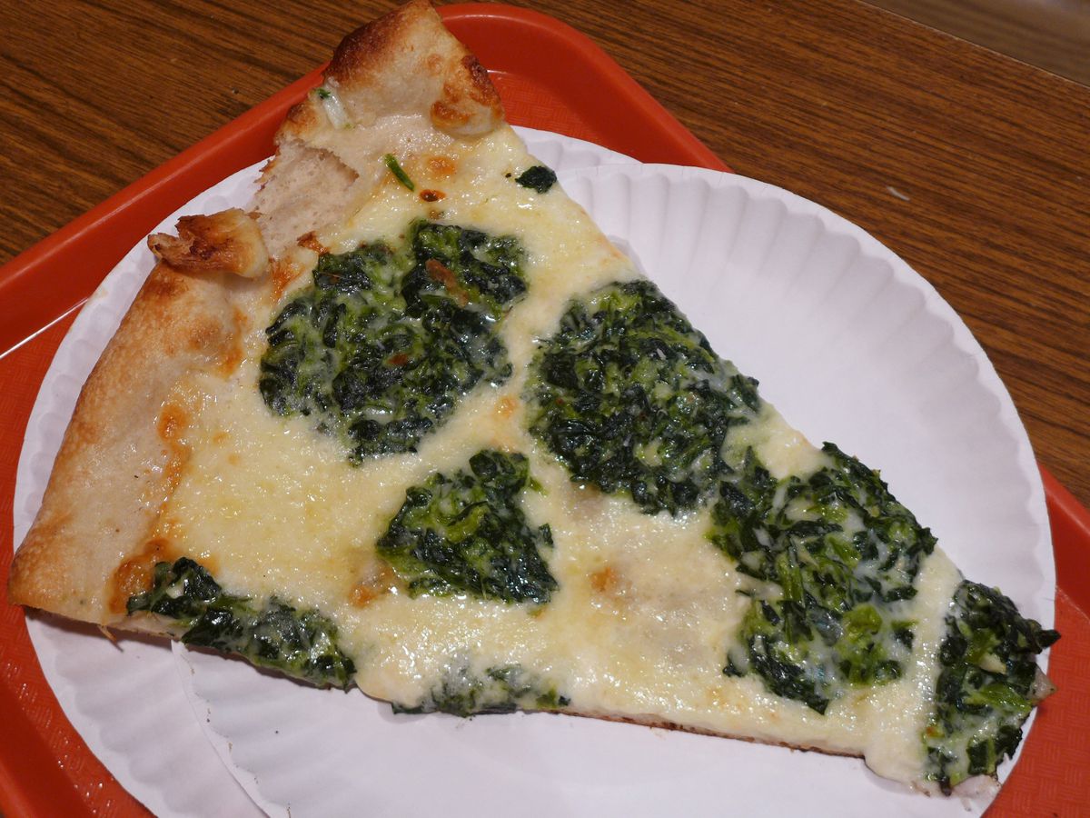 A wedge shaped slice of pizza with wads of green spinach on a white cheese background, with no tomato sauce.